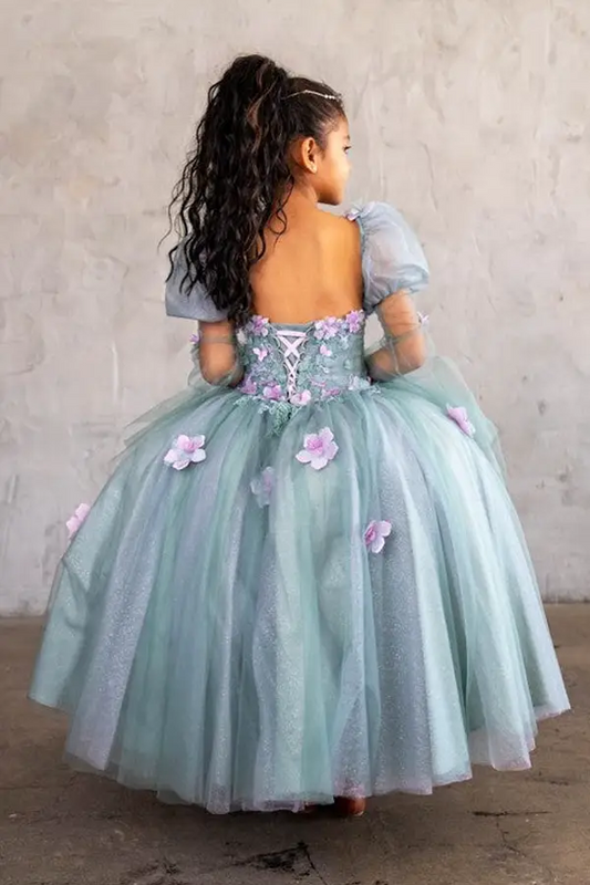 Young woman standing back-facing in sage ball gown with lace-up closure, 3D flower detailing, removable sleeves, and cascading skirt
