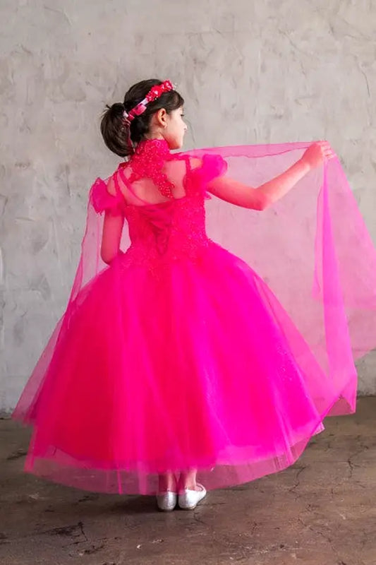 Young woman standing back-facing in fuchsia ball gown with lace-up closure, sequin bodice, dramatic cape, and cascading full skirt
