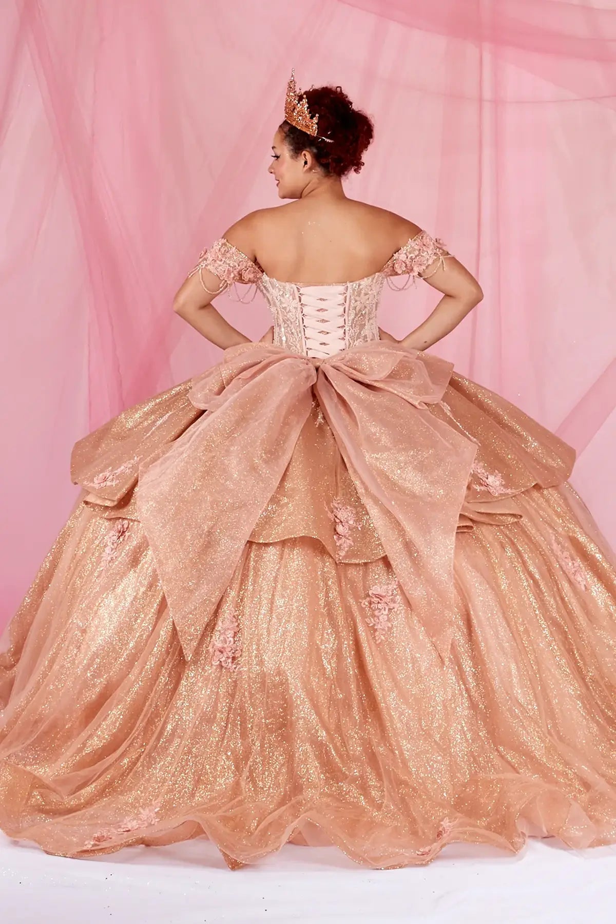 Back-Facing of a woman wearing Rose gold formal gown featuring lace-up back ruffled skirt overlay and chapel train