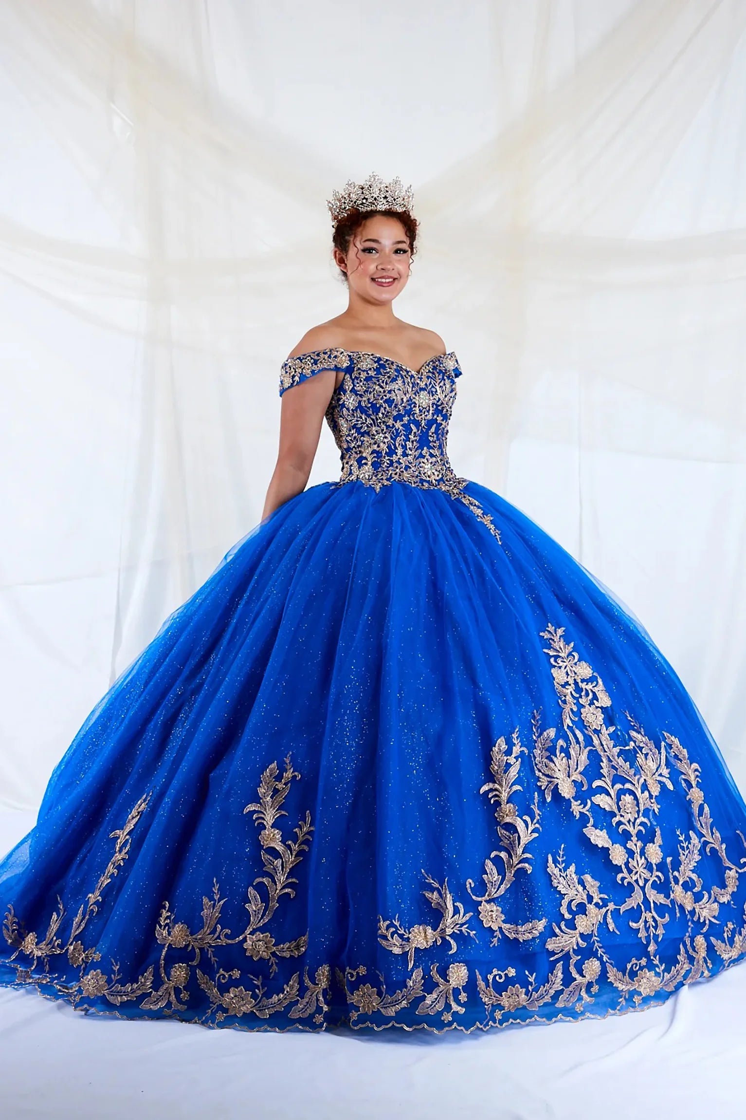 Front view of a woman wearing a royal blue quinceañera dress with embroidered lace appliques, off-shoulder A-line silhouette and glitter tulle overlay