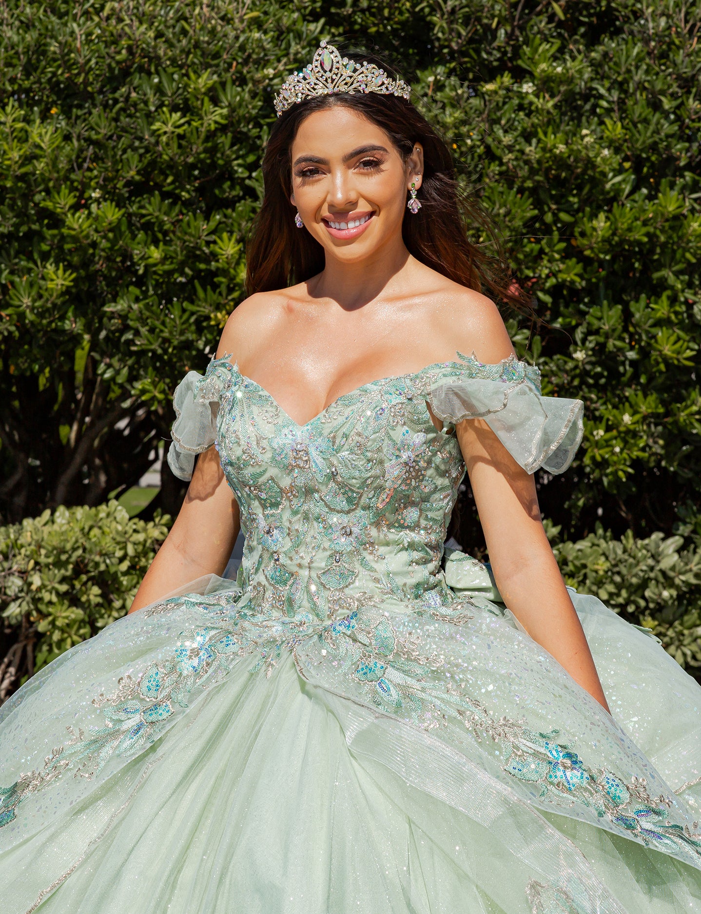 Girl in a light green, embellished dress with a tiara, standing outdoors with greenery in the background.