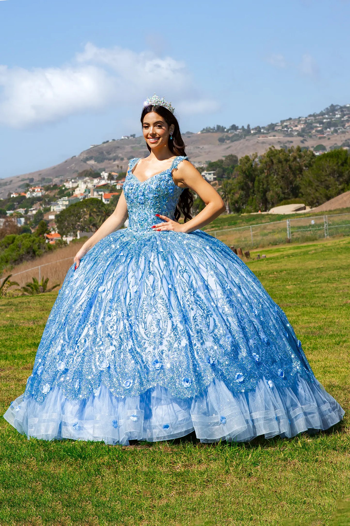 Woman in a blue quinceañera dress standing in a grassy field with hills in the background.